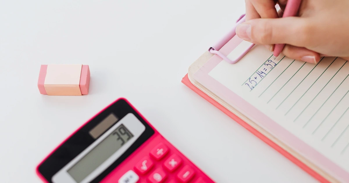 白色桌面上有粉色计算器、粉色橡皮和夹板笔记本，一只手正用粉色笔在纸上书写算式，计算器屏幕显示结果。